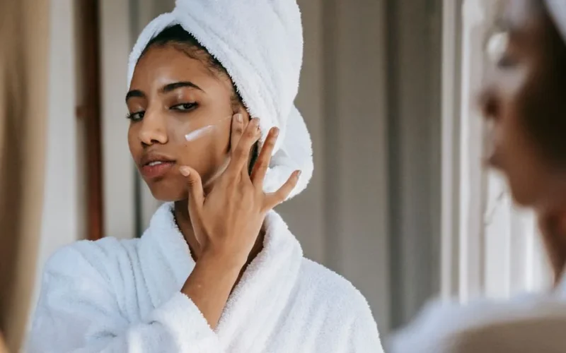 Crop attentive ethnic female in soft robe applying smear of moisturizing cream on cheek against mirror at home
