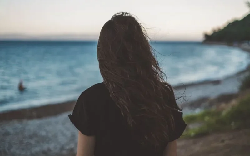 Woman with wavy hair standing by the serene seaside, facing the ocean at twilight.