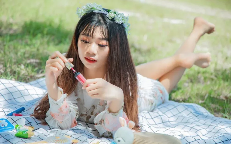A young woman with a floral crown applies lipstick while lying on a picnic blanket outdoors in a sunlit park.