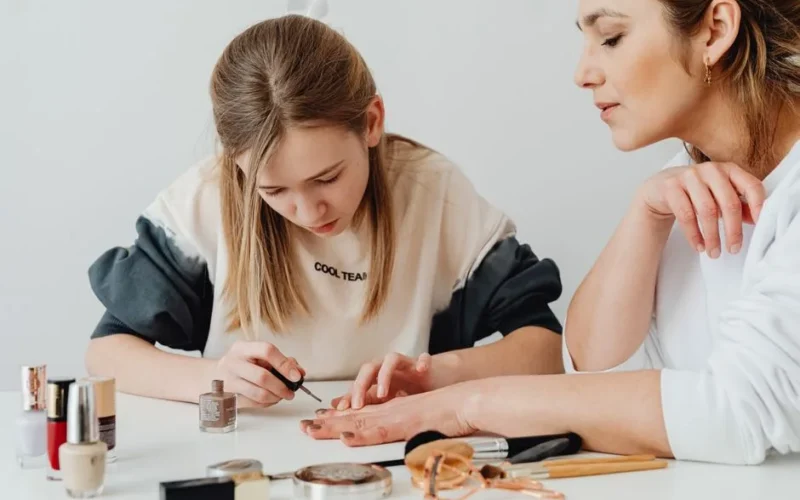 A mother and daughter enjoy quality time painting nails indoors, surrounded by cosmetics.