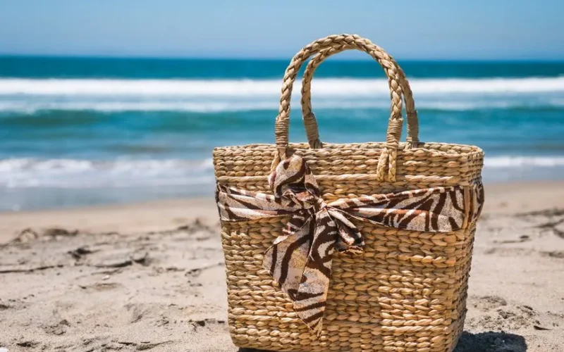 A stylish woven beach bag sits on a sunny beach with waves in the background.
