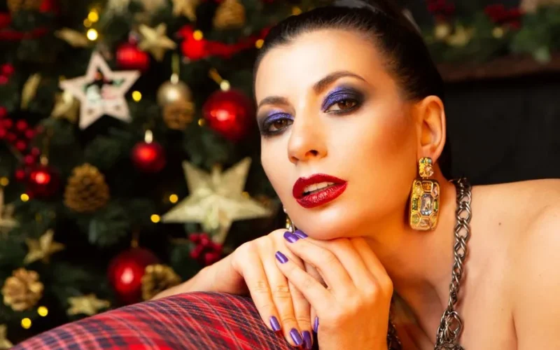 A glamorous woman poses with festive makeup by a Christmas tree.