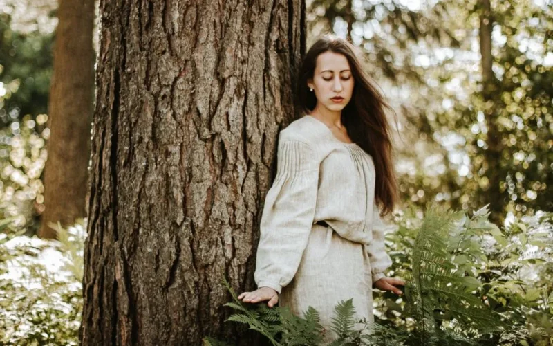 Woman in linen dress posing by tree in a serene forest setting.