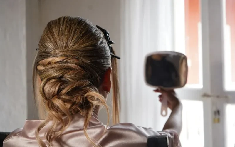 A woman with an elegant braided updo admires her hairstyle in a hand mirror near a window.