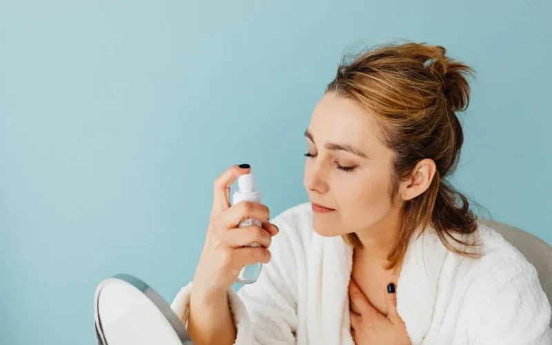 Woman in a bathrobe sprays skincare product onto her face with a blue background.