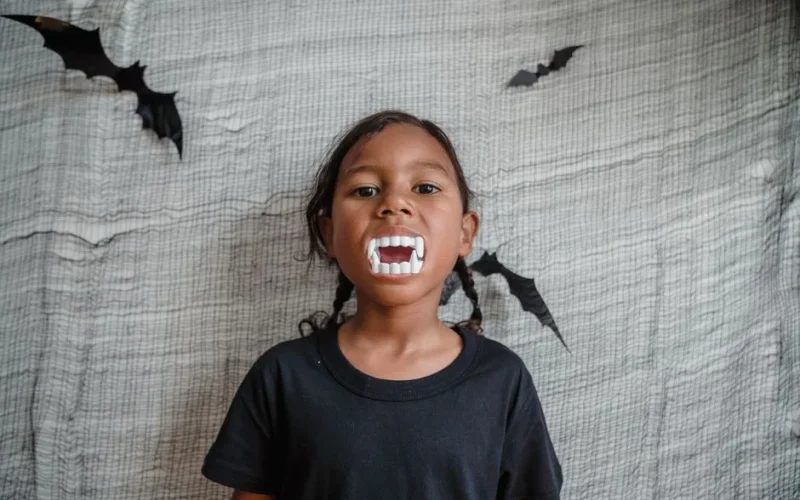 A young girl in a black t-shirt showing vampire teeth decorations during Halloween.