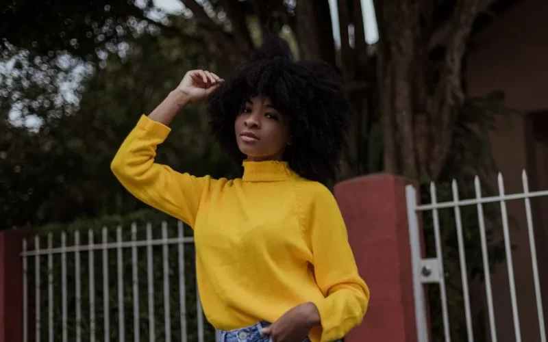 Low angle of black female with Afro hairstyle wearing vivid yellow sweater touching hair while standing on street