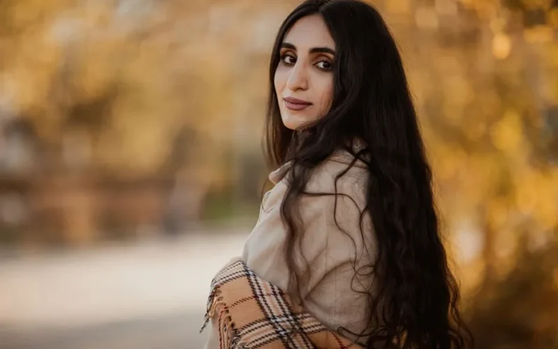 A woman with long dark hair looking over her shoulder amidst autumn foliage.