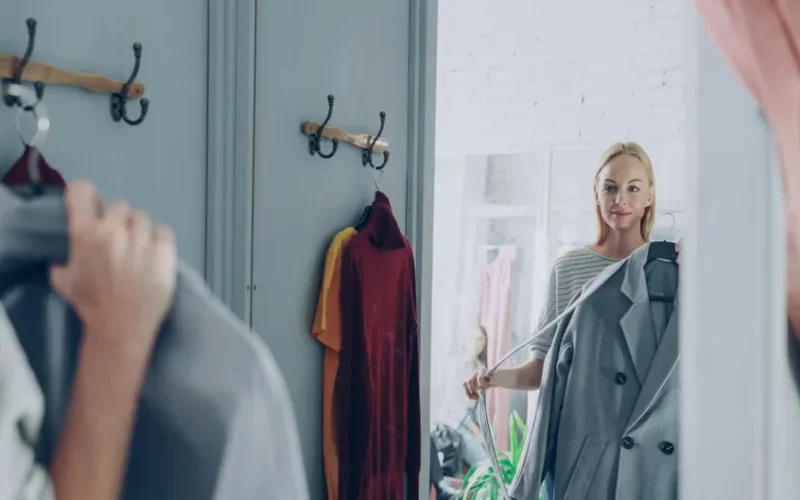 Woman holding a jacket, reflecting in a store mirror, deciding on her outfit.