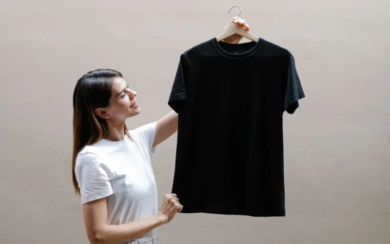Side view of self assured young woman with long dark hair in stylish outfit smiling while holding hanger with black t shirt against gray wall