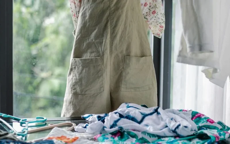 Crop anonymous woman in casual wear sorting and picking garments from clothes heaped on table in light cloakroom