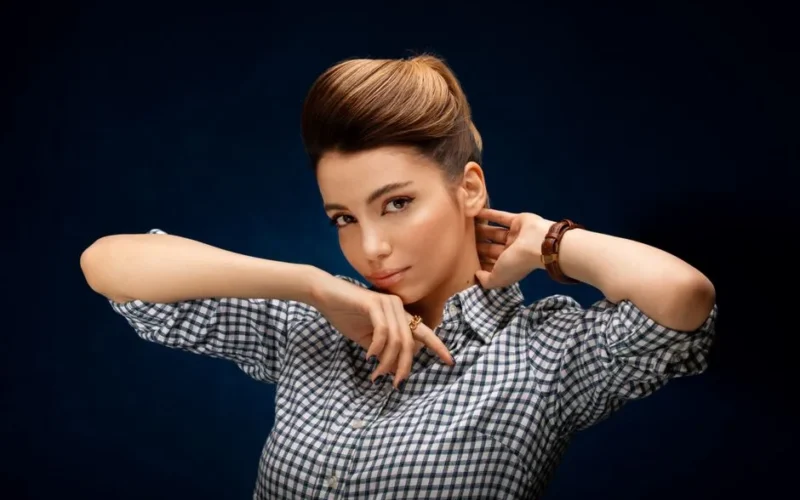 Stylish young woman with hairdo touching neck and chin while looking at camera with deep gaze