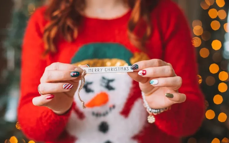 Close-up of hands holding a Merry Christmas ribbon, wearing a festive red sweater with cheerful nail art.