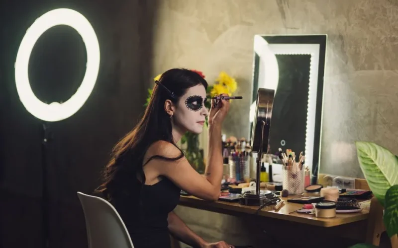 A woman in a black dress applies Halloween makeup by a mirror with cosmetics and a ring light.