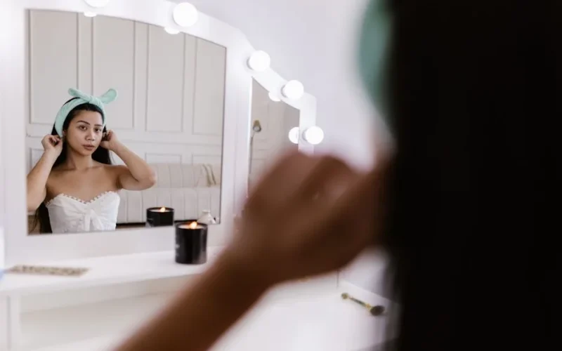 Young woman adjusts hairband at vanity mirror with candles, in serene home setting.
