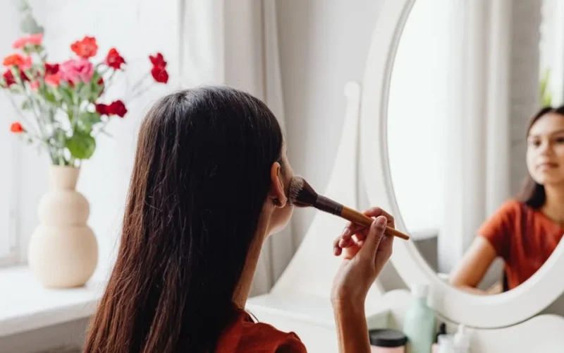 A woman applying makeup with a brush while facing a mirror indoors.