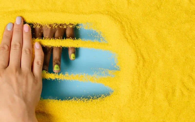 Creative composition of hands with colorful nails in vibrant yellow sand. Perfect for fashion or art concepts.