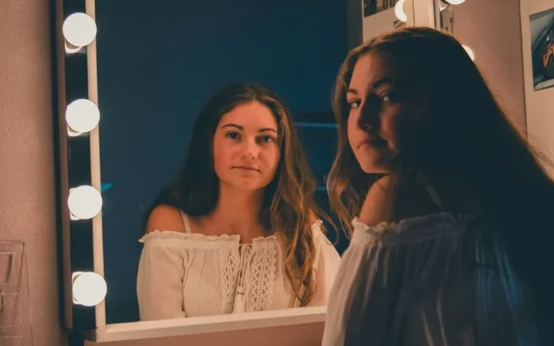 A young woman gazes thoughtfully into a vanity mirror in a softly lit room.