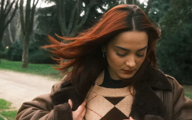 Portrait of a woman in a stylish brown coat and sweater, standing outside in a park setting.