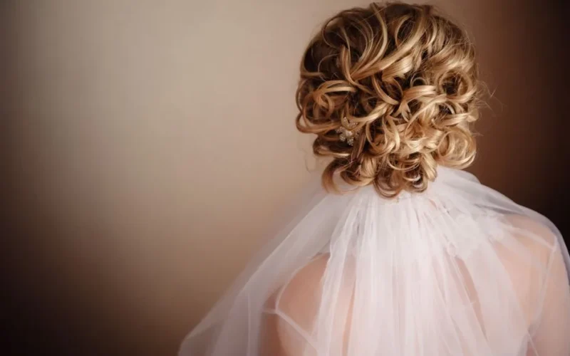 A beautiful bridal hairstyle featuring an elegant curly blonde updo accented with a white veil.