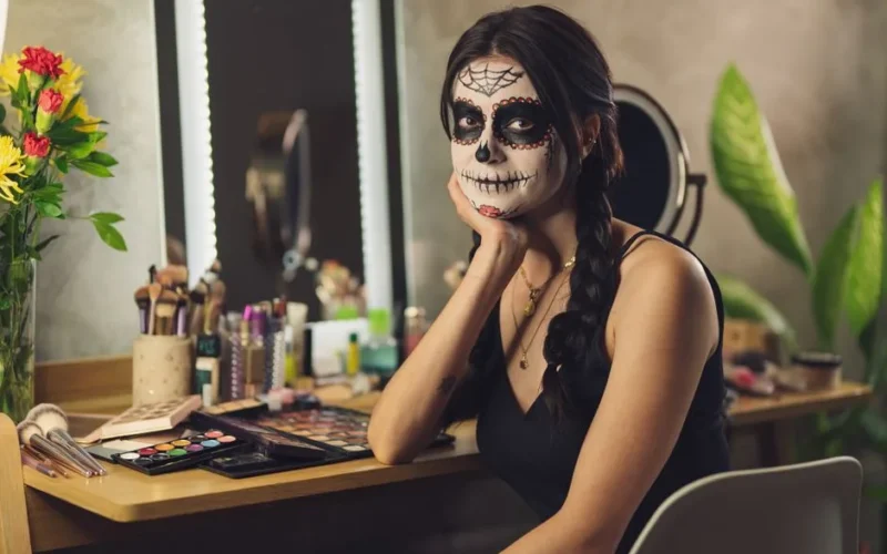A woman with Catrina face paint sits in a well-lit vanity, capturing the essence of Dia de los Muertos.