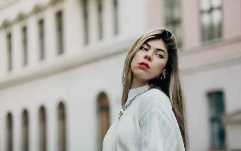 Stylish young woman in a white sweater poses confidently in a modern urban environment.