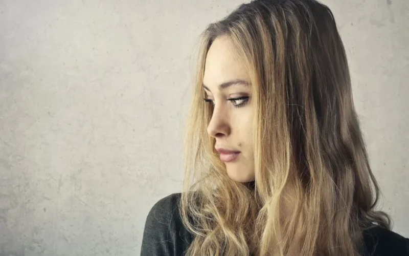 A pensive young woman with long blonde hair looking to the side against a neutral background.