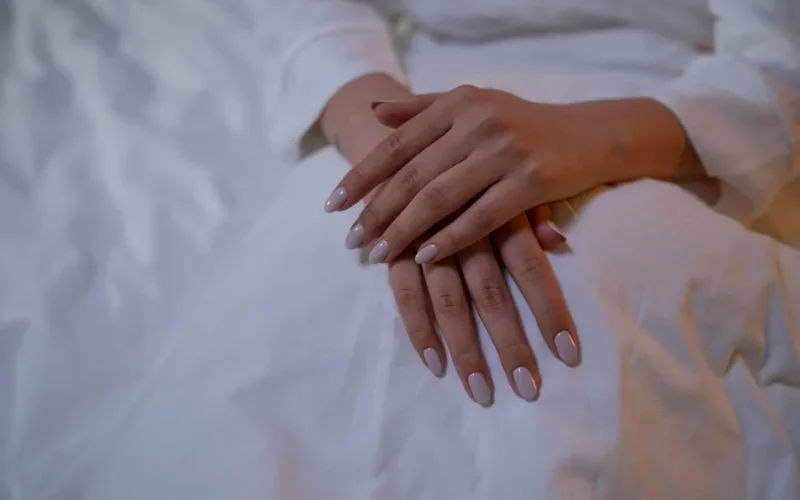 Close-up of hands resting, showcasing elegant nude nails on a white fabric.