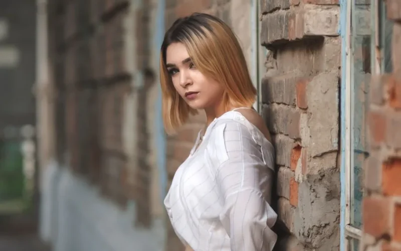 Model with blonde hair in white top leaning against a brick wall, showcasing modern fashion.