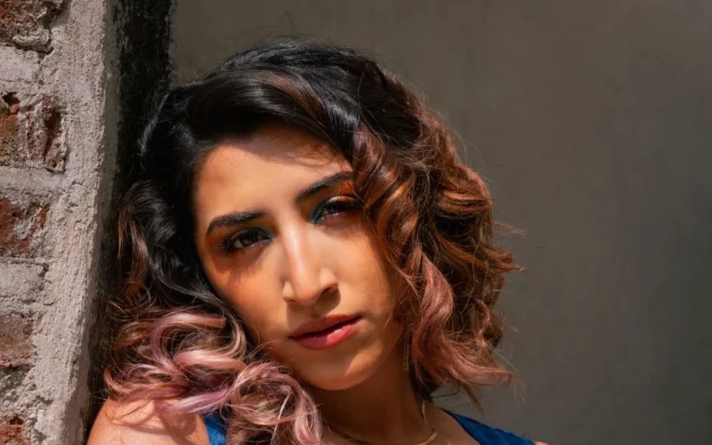 Close-up portrait of a woman with curly hair, highlighted by natural sunlight against a rustic brick wall.