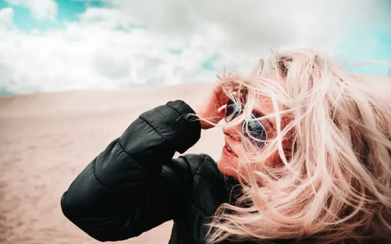 A woman with windswept hair in sunglasses enjoys a breezy day on a sandy beach.