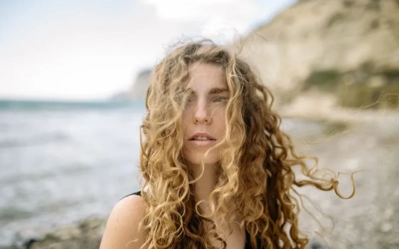 Close-up portrait of a woman with curly hair on a sandy beach with a blurred background.