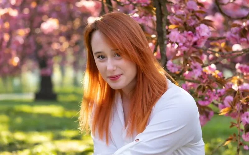 Portrait of a woman with red hair smiling among pink cherry blossoms in spring.