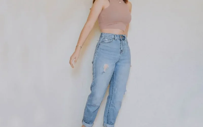 Adult in casual wear with crop top and ripped jeans, standing indoors against a neutral wall.