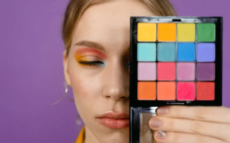 Close-up of a woman with colorful eyeshadow and palette against a purple background.