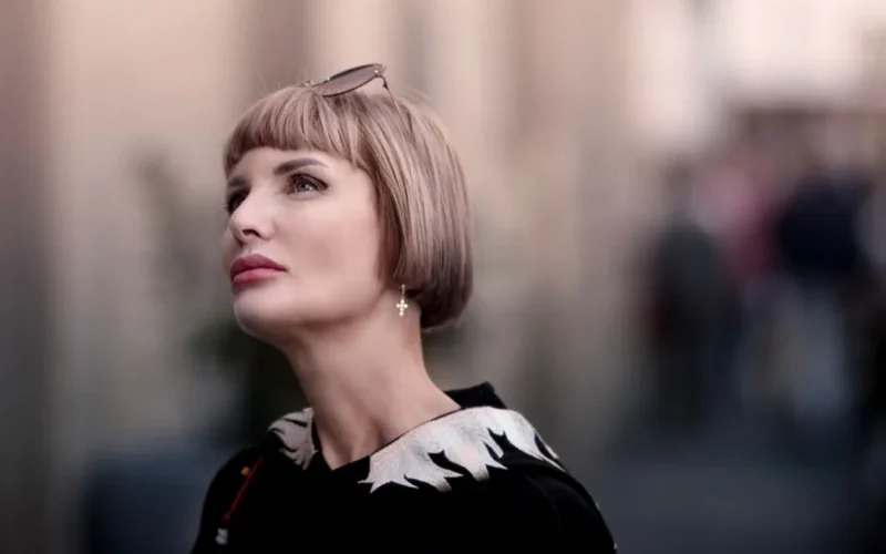 Stylish portrait of a woman with blonde bob haircut and sunglasses, looking up with a blurred urban background.