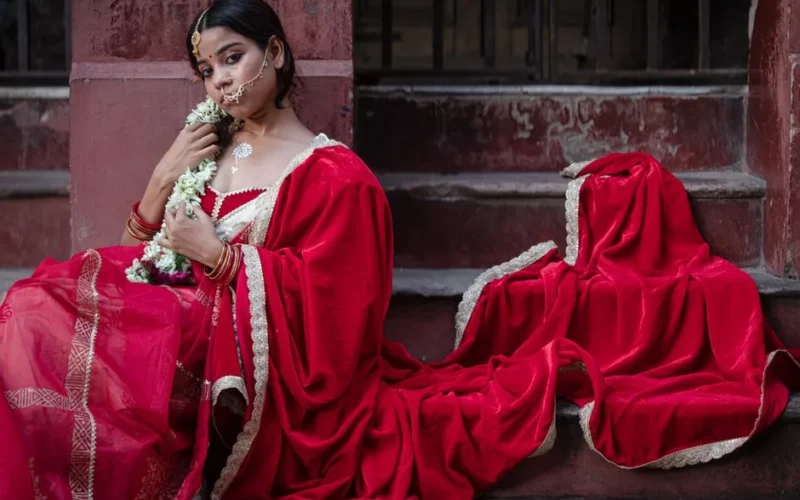 A woman in a vibrant red sari, adorned with traditional Indian jewelry, seated outdoors.
