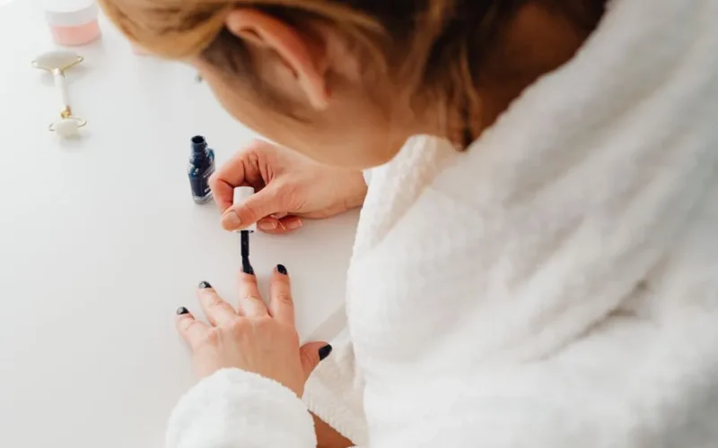 A woman in a bathrobe paints her nails indoors as part of a self-care routine.