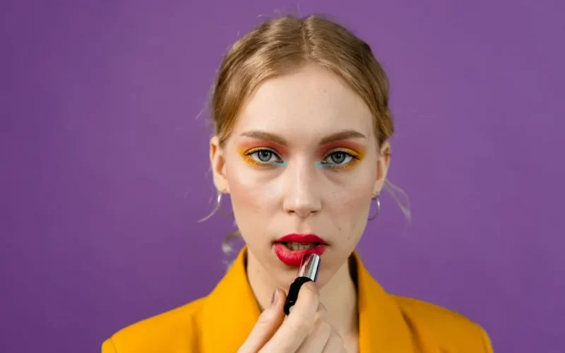 Close-up portrait of a woman applying red lipstick with colorful eye makeup against a purple background.