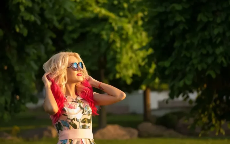 A stylish woman with colorful hair enjoys a sunny day in a park, capturing the joy of summer.