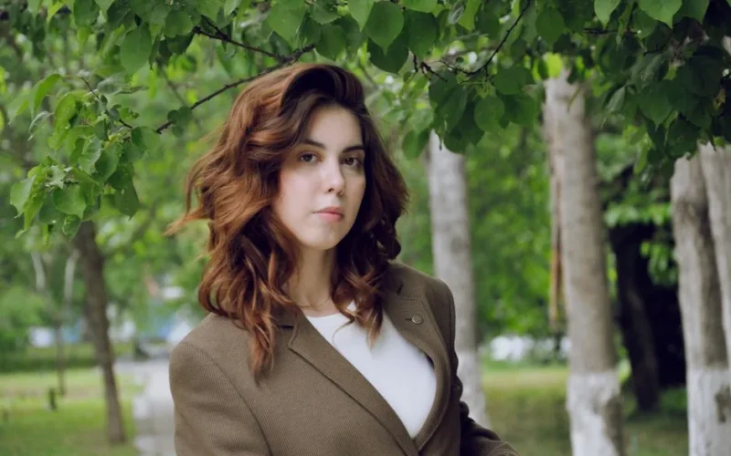 Woman with wavy hair wearing a brown blazer, standing confidently in a lush green outdoor setting.