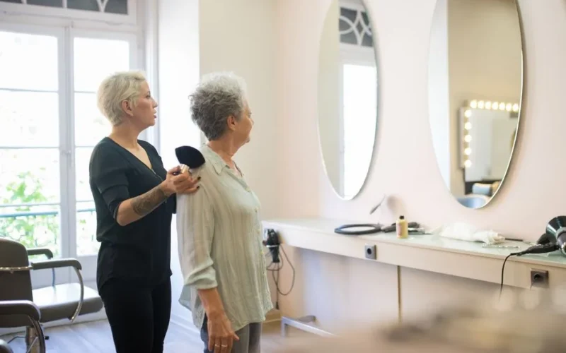 Hair stylist assisting a senior client in a modern salon with large mirrors.