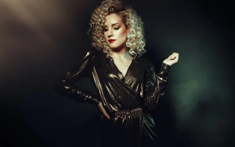 Stylish woman with curly blond hair posing in a shimmering outfit against a dark studio backdrop.