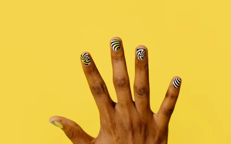 Close-up of a hand with vibrant patterned nail art against a bright yellow background.