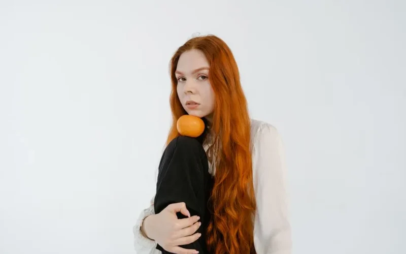 A woman with long red hair poses with an orange against a plain white background.