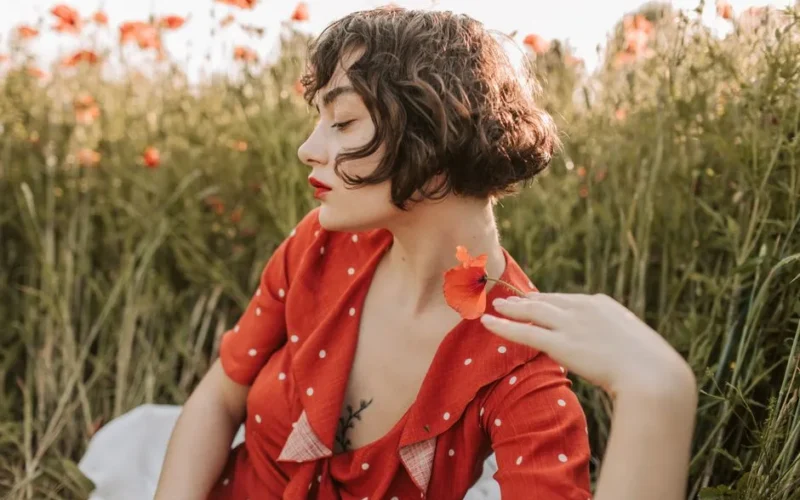 Brunette woman in red polka-dot dress posing in a flower field with red lips and short hair.