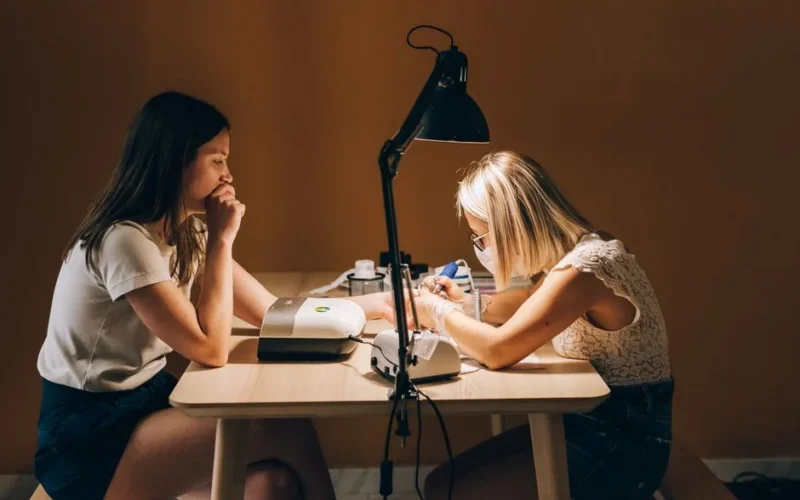 A manicurist wearing a face mask focuses on a customer's nails in a dimly lit room.