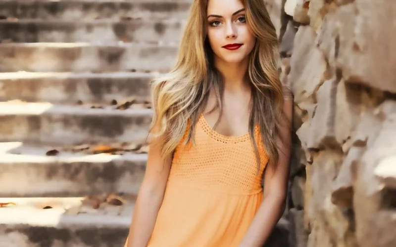 A beautiful woman with long blonde hair and red lips posing by a stone wall outdoors.