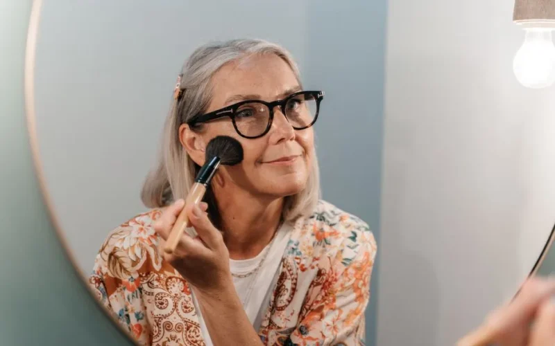 Senior woman with glasses applying makeup while looking in a mirror, reflecting a joyful moment.