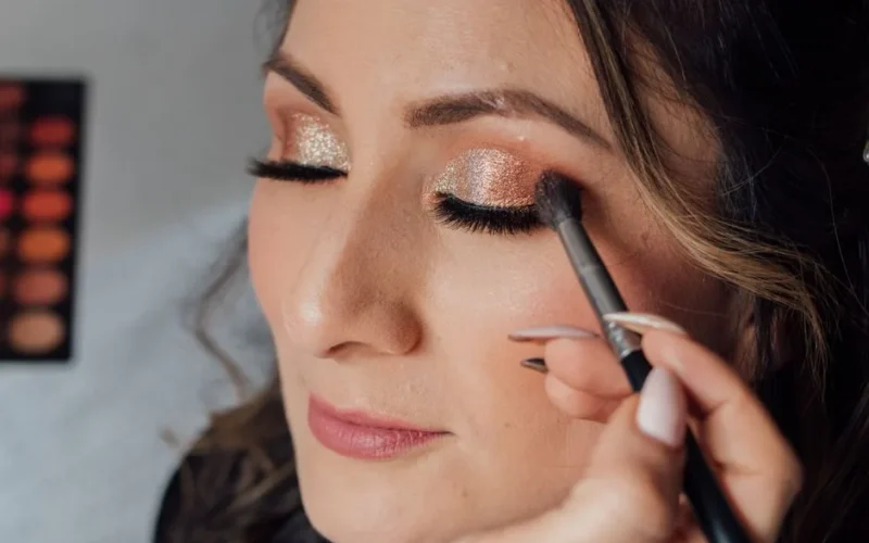 Woman receiving glamorous makeup with glitter eyeshadow and long lashes.